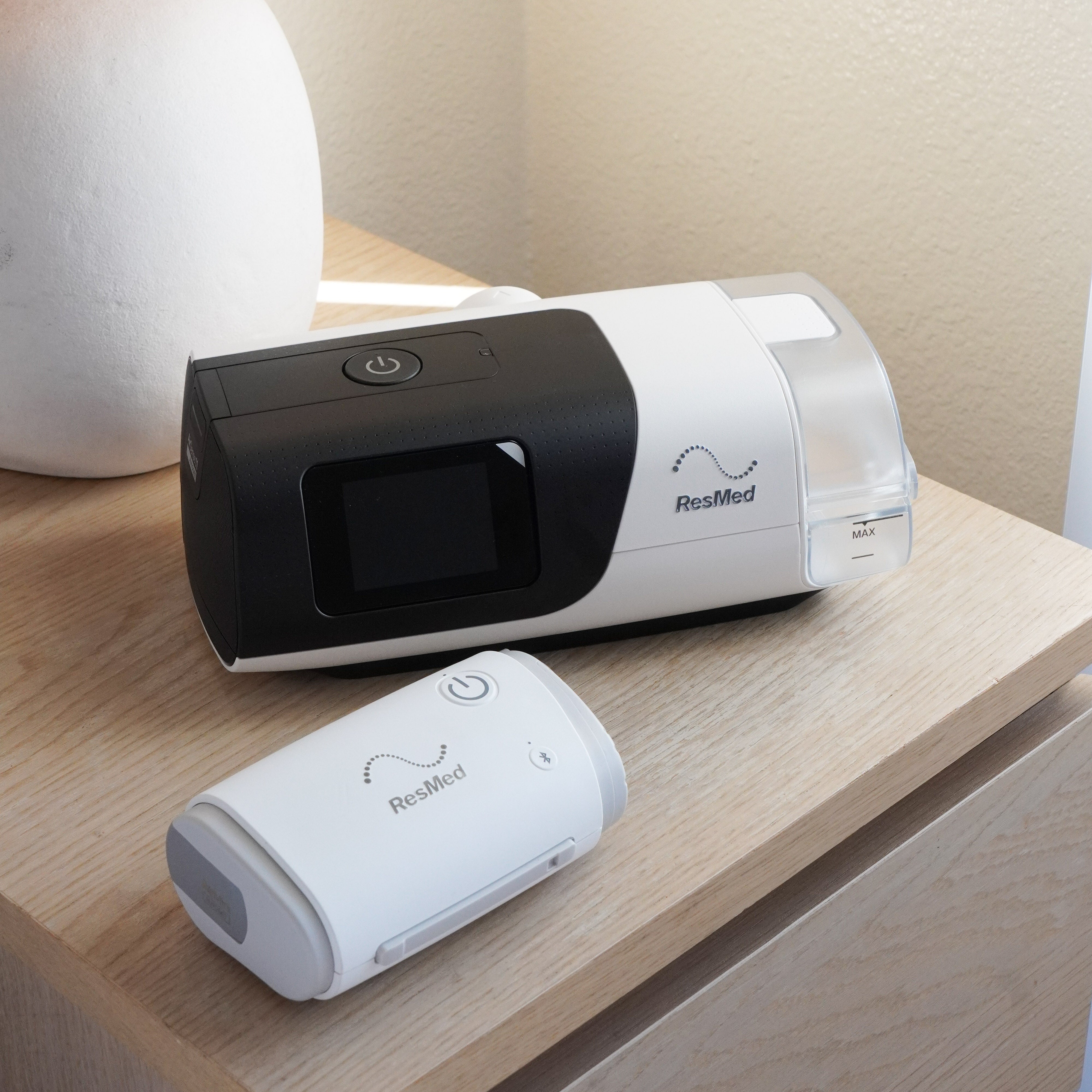 The ResMed AirMini next to the ResMed AirSense 11 on a bedside table for size comparison. The ResMed AirMini is much smaller due to its compact travel size.